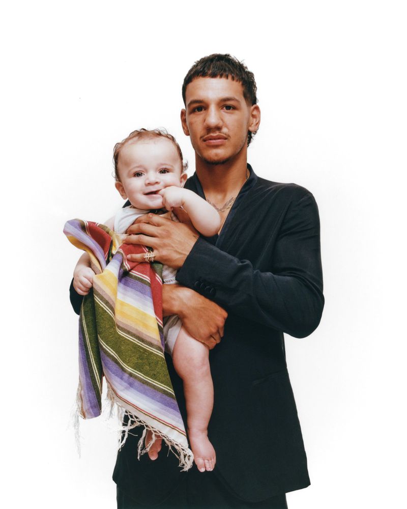 Studio shot of an adult male in a black jacket holding a baby wrapped in a colorful striped blanket, both looking at the camera.