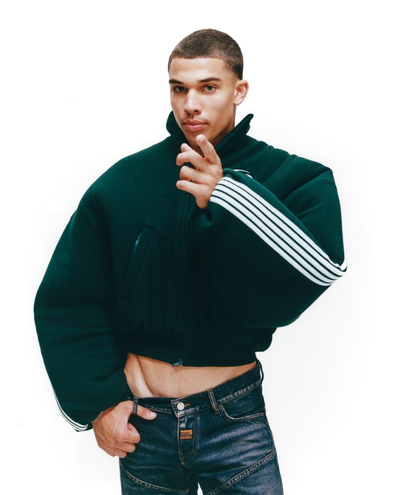 A lean adult male poses in a studio wearing a green jacket with white stripes and jeans, pointing his finger towards the camera.
