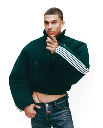 A lean adult male poses in a studio wearing a green jacket with white stripes and jeans, pointing his finger towards the camera.