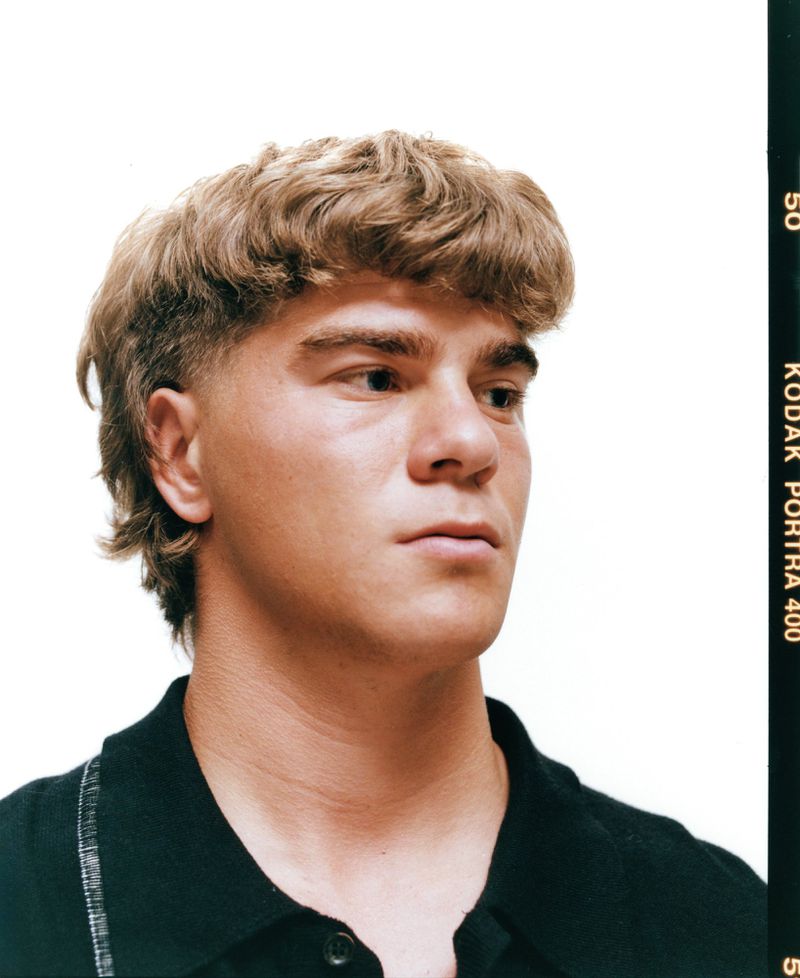 Portrait of a young fair-skinned man with a blonde mullet hairstyle, wearing a black shirt, against a bright white backdrop, shot on film.