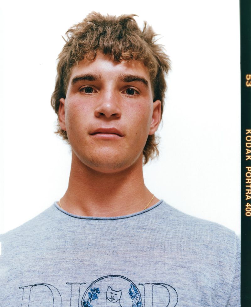 Close-up portrait of a young adult male with tousled hair wearing a gray T-shirt with a graphic, against a plain white backdrop in a photo studio.