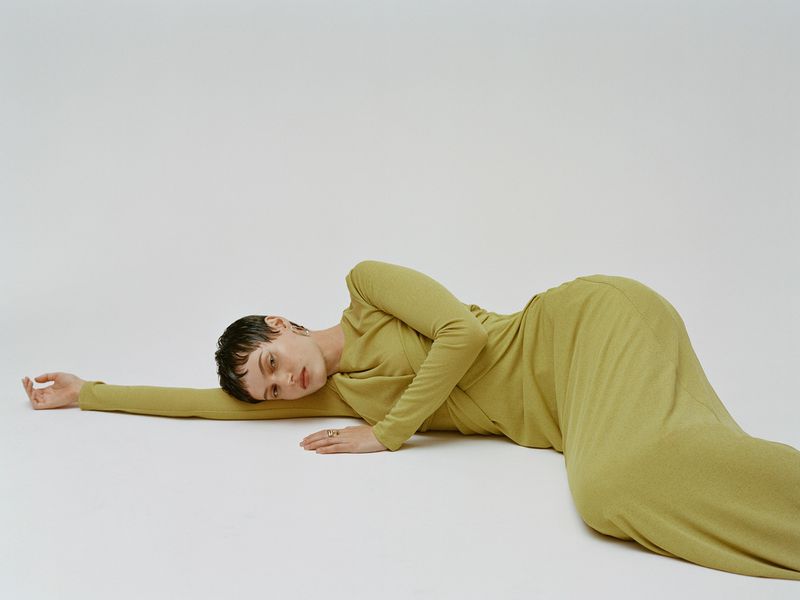 Adult woman in a long-sleeved chartreuse dress lies on her side on a plain white surface in a studio setting with soft lighting.