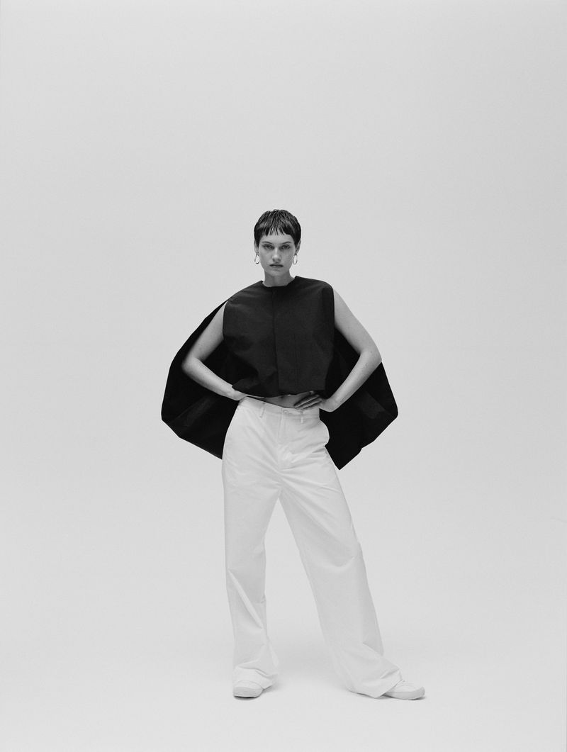 Black and white studio shot of an adult female fashion model posing in a dark, sleeveless top and white wide-leg pants with hands on her waist.