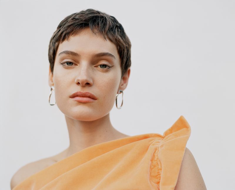 Close-up portrait of model Greta Hofer wearing a peach top and silver earrings against a white background.