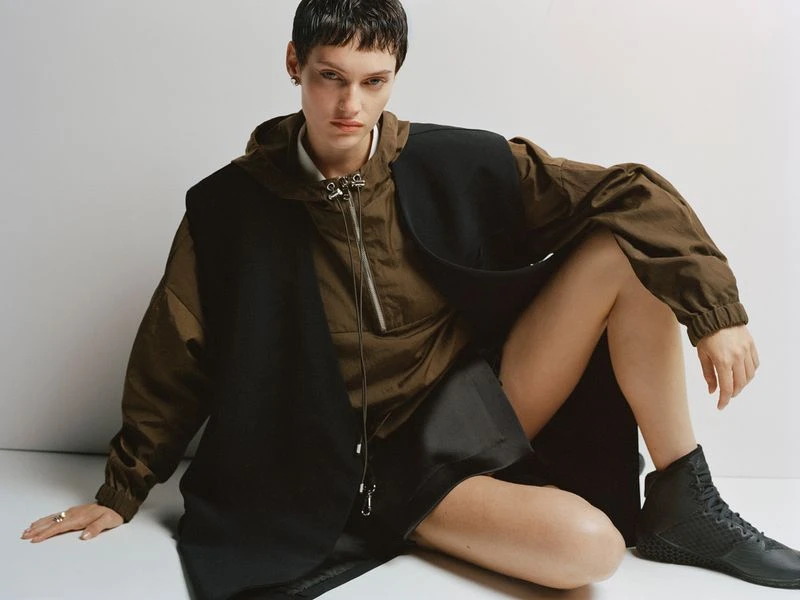 A woman with short dark hair sits on a white surface wearing an olive green hooded jacket, black vest, and black shoes looking at the camera.