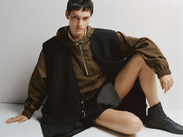 A woman with short dark hair sits on a white surface wearing an olive green hooded jacket, black vest, and black shoes looking at the camera.