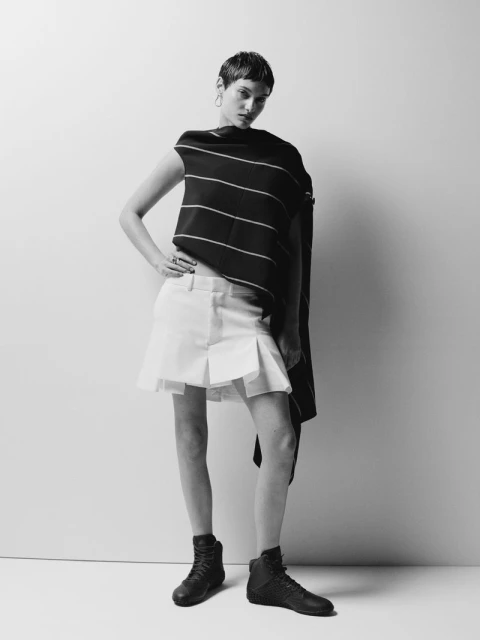Black and white fashion shot of a woman in a striped top, white skirt, and black boots, posing in a studio against a white background.