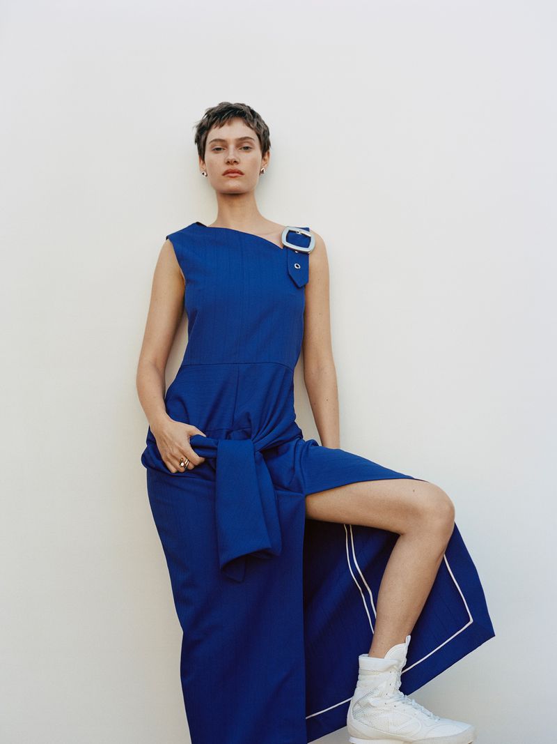 Fashion shot of a woman in a blue dress with a leg extended, wearing white sneakers against a white background. Studio lighting.