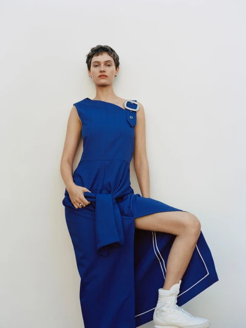 Fashion shot of a woman in a blue dress with a leg extended, wearing white sneakers against a white background. Studio lighting.