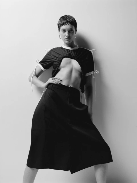 Black-and-white fashion shot of a young adult woman posing in a cropped top and skirt, set against a simple studio backdrop.