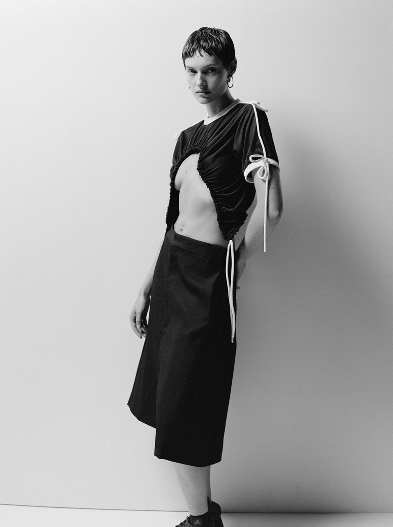 A woman poses in a black top and skirt against a light wall, showcasing a modern fashion look in a studio setting.