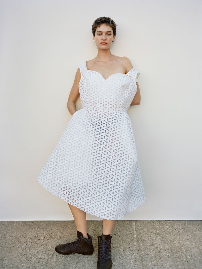 Elegant woman in a stylish white openwork dress poses against a neutral background, complemented by unique dark footwear for a fashion statement.