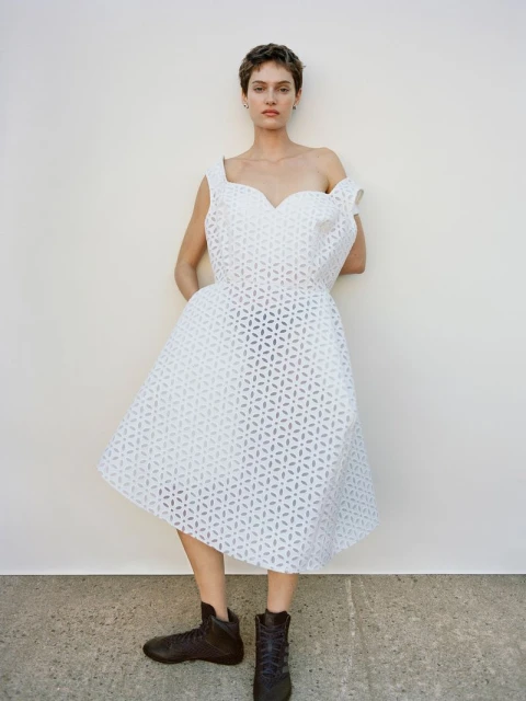 Elegant woman in a stylish white openwork dress poses against a neutral background, complemented by unique dark footwear for a fashion statement.