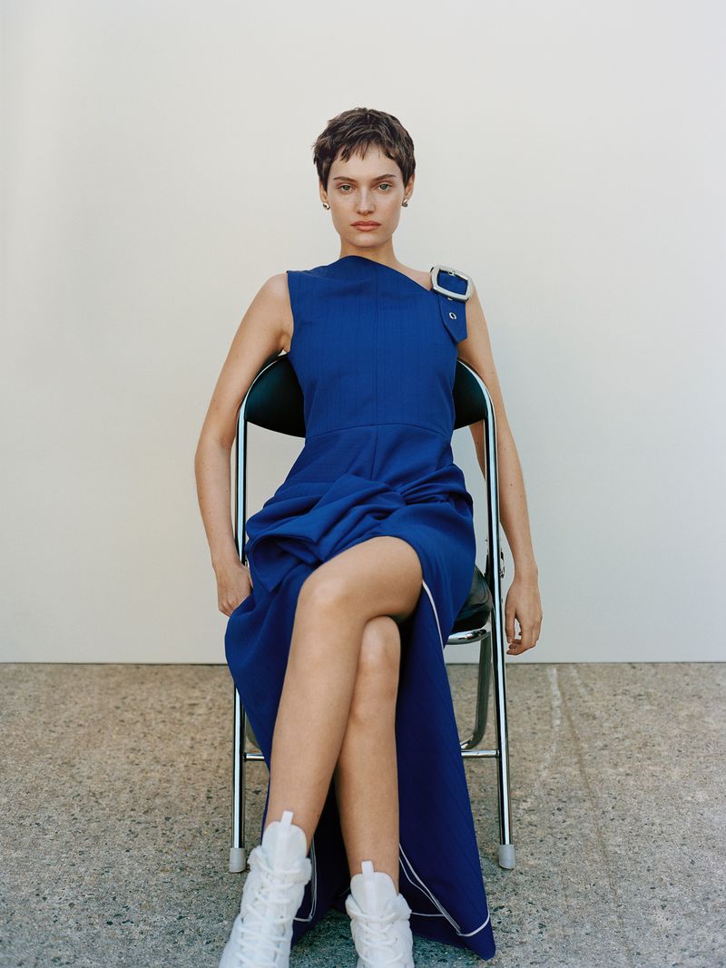 Model with short brown hair wearing a blue dress seated on a metal chair against a plain white background