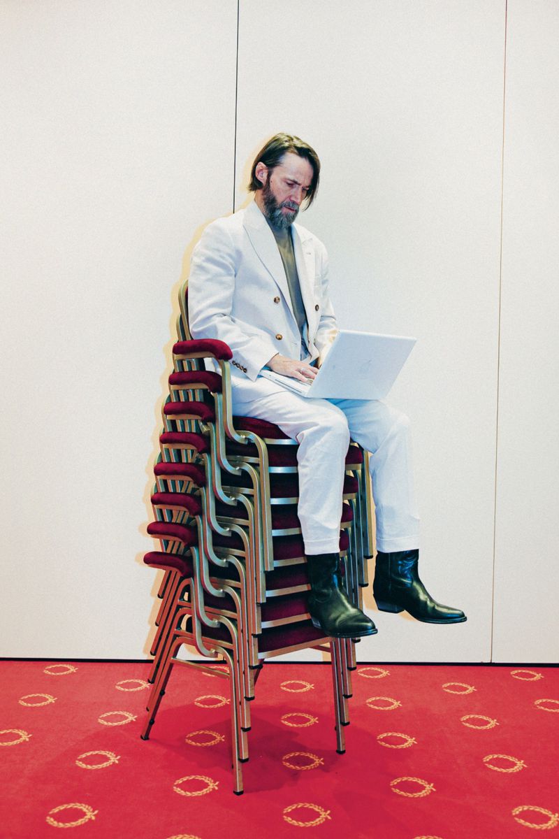 Adult male sits on a stack of red chairs and uses a laptop computer in an indoor setting with a red patterned floor.