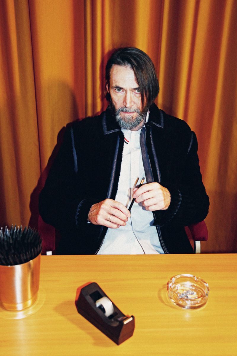 Man with beard in black jacket sitting at a desk with pens, tape dispenser, and ashtray against a gold curtain backdrop.
