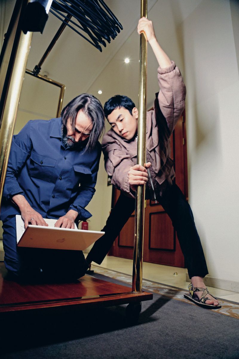 Two men in an interior setting, one kneeling by a laptop on a luggage cart and another gripping a vertical brass pole.