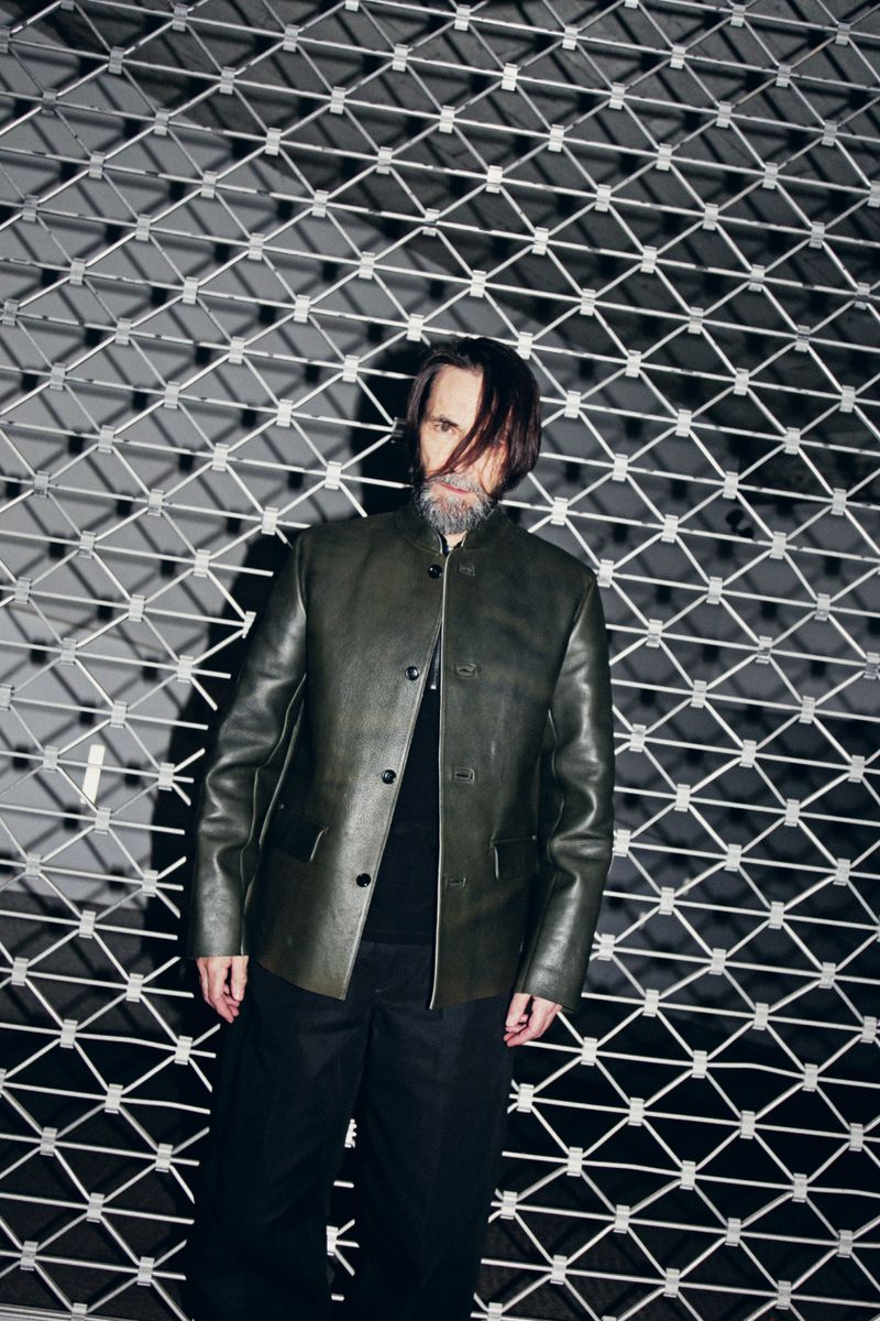 Adult male in dark jacket and pants standing against a metal gate. He has long dark hair and a salt-and-pepper beard. The background is dark.
