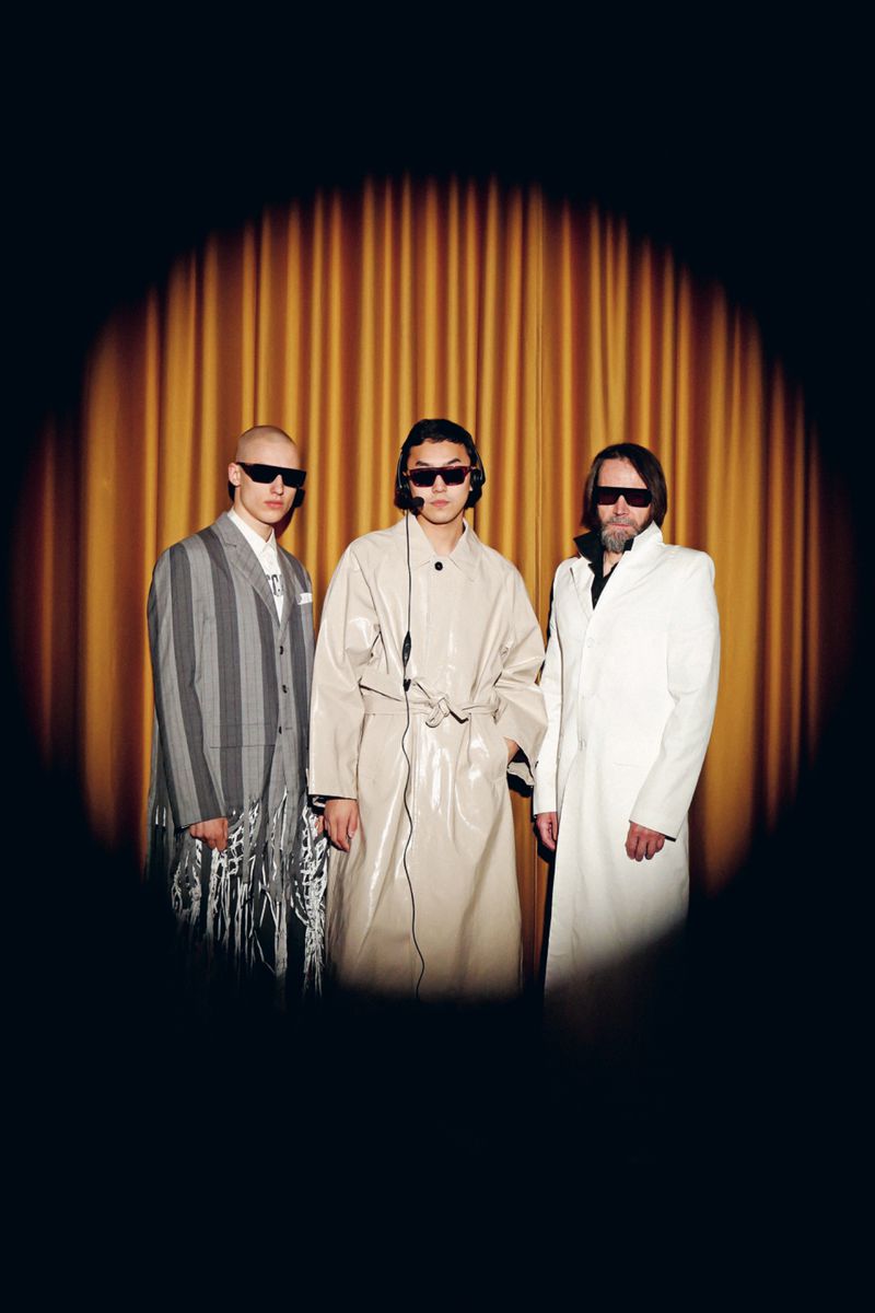 Three adult male models in fashionable coats and sunglasses stand in front of a gold curtain with a spotlight effect in a studio setting.