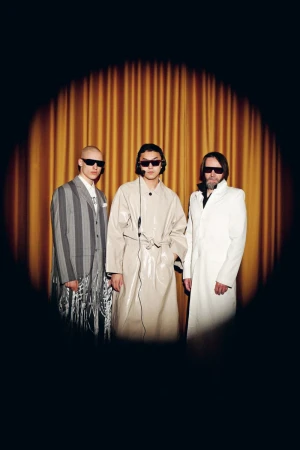 Three adult male models in fashionable coats and sunglasses stand in front of a gold curtain with a spotlight effect in a studio setting.