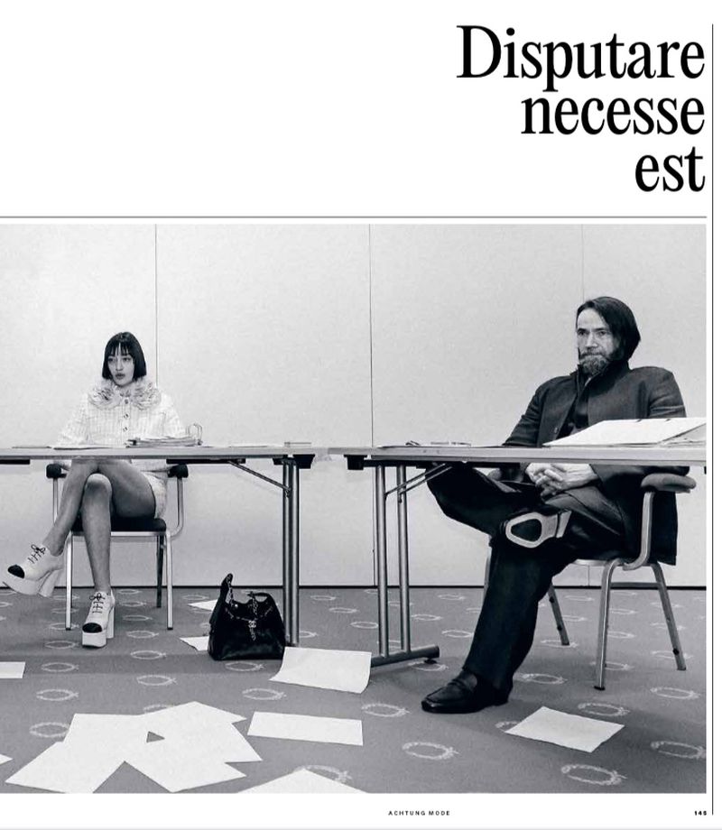 Editorial fashion shot in black and white featuring an adult female and male seated at tables with scattered papers and a handbag.