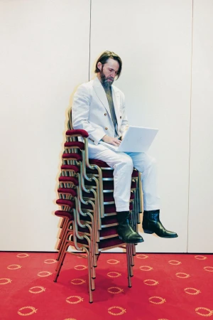 Man wearing a white suit sitting on a towering stack of chairs while using a laptop
