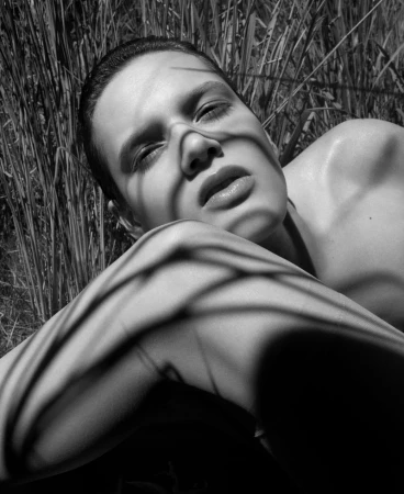 Close-up portrait of a model lying outdoors amidst tall grass with stark shadow patterns cast across the face.