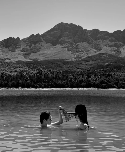 Black and white shot of two people waist-deep in water, a net between them, with a tree line, water, and a large mountain backdrop.