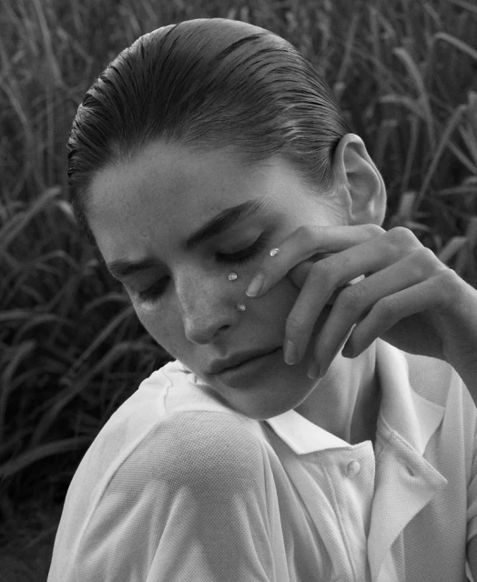 Monochrome portrait of a young woman with wet slicked-back hair, white collared shirt, and teardrop embellishments on her cheek in tall grass.