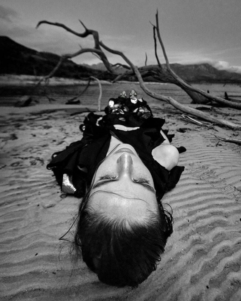 Monochromatic portrait of a woman lying on a sandy beach with closed eyes, featuring driftwood and distant mountains under a cloudy sky.