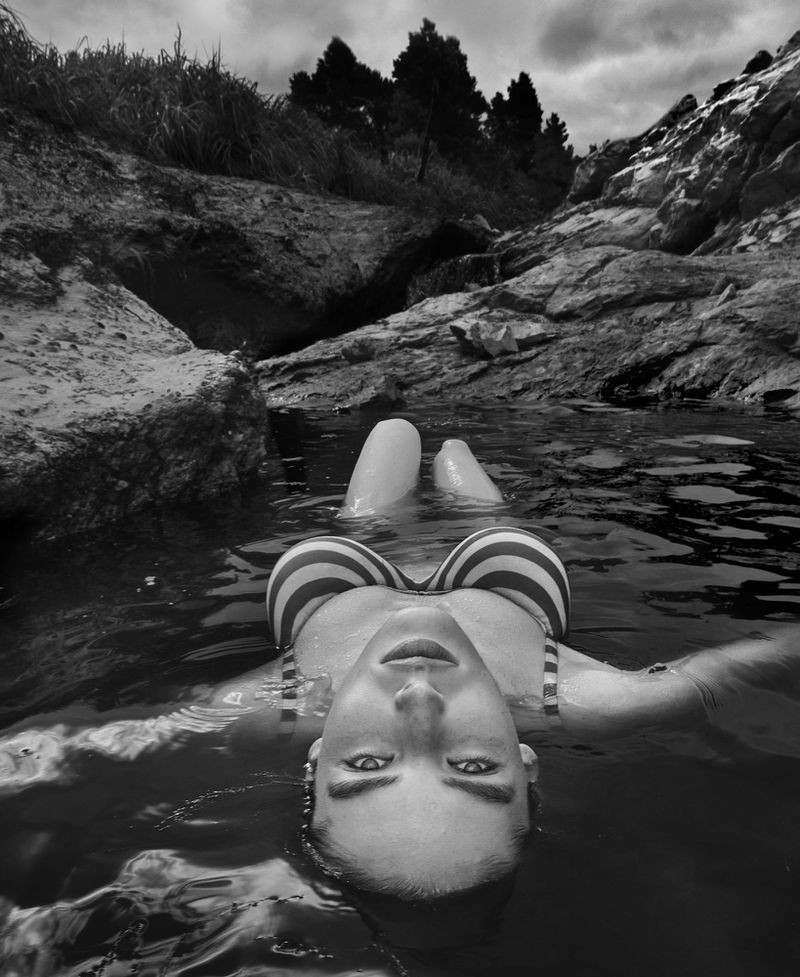 Black and white portrait of a woman in a striped bikini floating in water among rocks and trees, looking directly at the camera.