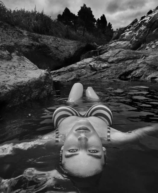 Black and white portrait of a woman in a striped bikini floating in water among rocks and trees, looking directly at the camera.