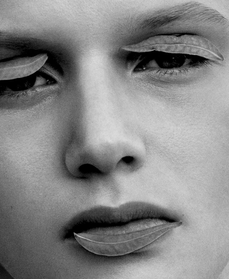 Black and white close-up of a woman's face with leaves covering her eyes and mouth, creating a surreal and artistic effect.