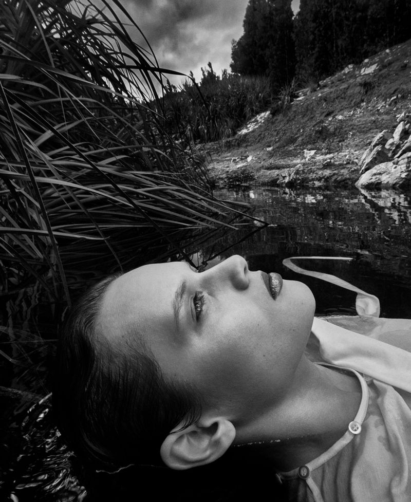 Monochrome close-up of a woman lying in water surrounded by tall grass, reflecting cloudy skies in a serene outdoor nature scene.