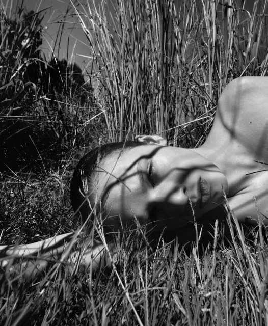 Black and white image of a female lying in a field of tall grass, face towards the sky, with grass shadows across her face.