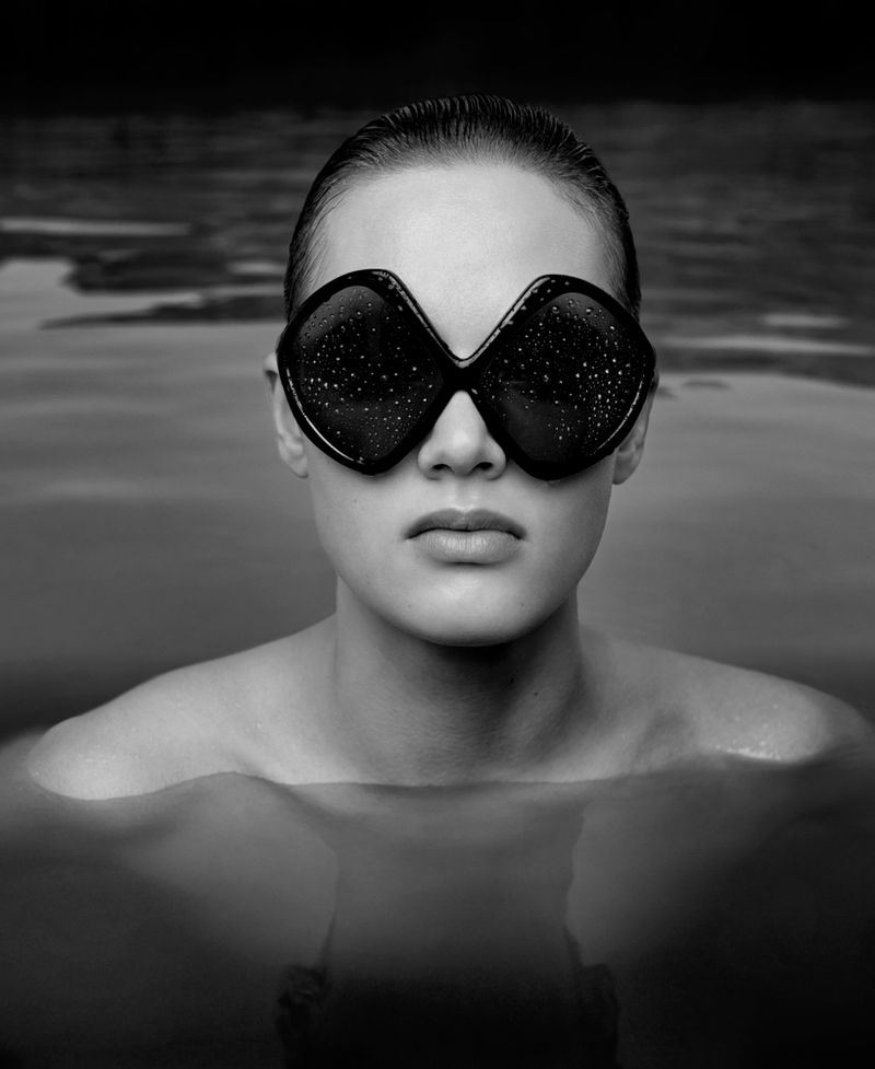 Monochromatic portrait of a woman in water wearing oversized sunglasses with a wet slicked-back hairstyle, focusing on her face.