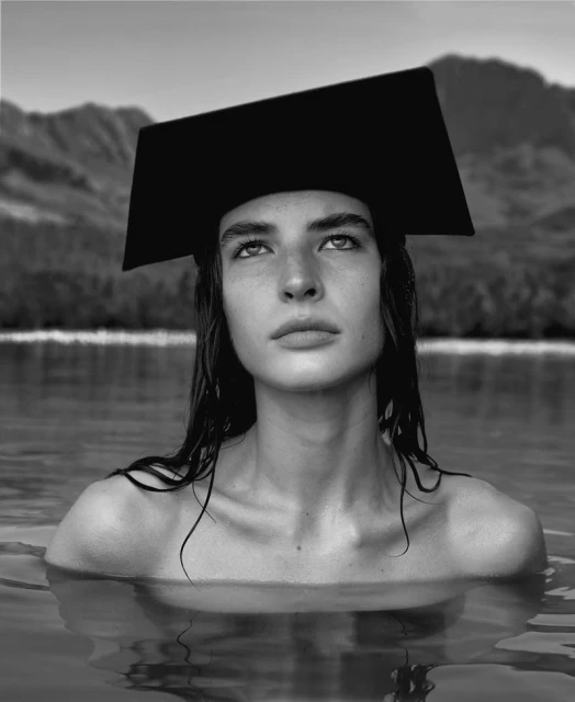 Monochromatic portrait of a woman in water with a square hat, mountains and lake visible in background, creating a serene and artistic scene.