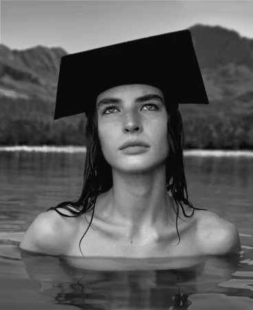 Model wearing a structured black hat while submerged in water with mountains and water visible in background