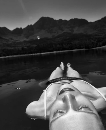 Woman floating on her back in a lake with mountains in the background, photographed in black and white with visible light reflections.