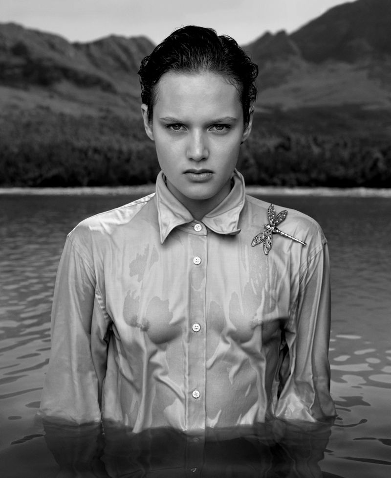 Monochromatic portrait of a young woman in water, wearing a wet button-down shirt and a dragonfly brooch, with a mountain backdrop.