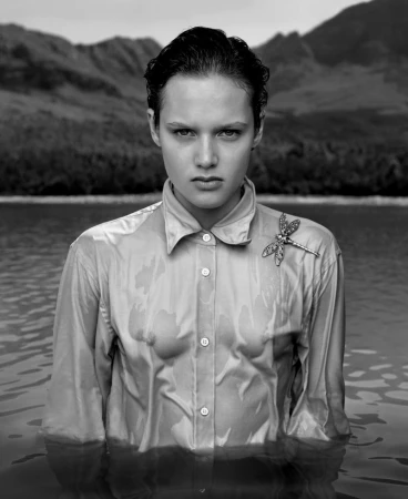 Monochromatic portrait of a young woman in water, wearing a wet button-down shirt and a dragonfly brooch, with a mountain backdrop.
