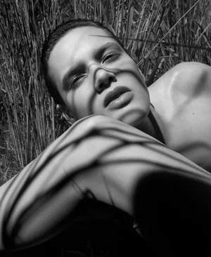 Close-up portrait of a model lying outdoors amidst tall grass with stark shadow patterns cast across the face.