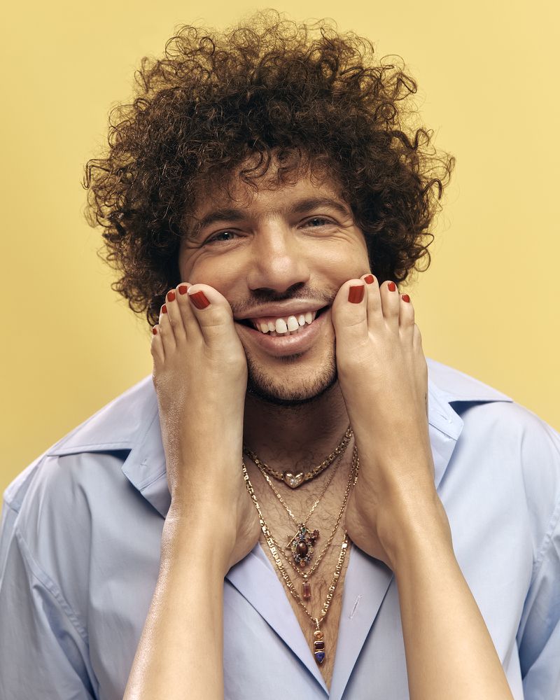 A man with curly hair smiles as a pair of feet with red toenails frame his face against a plain yellow background in a studio shot.