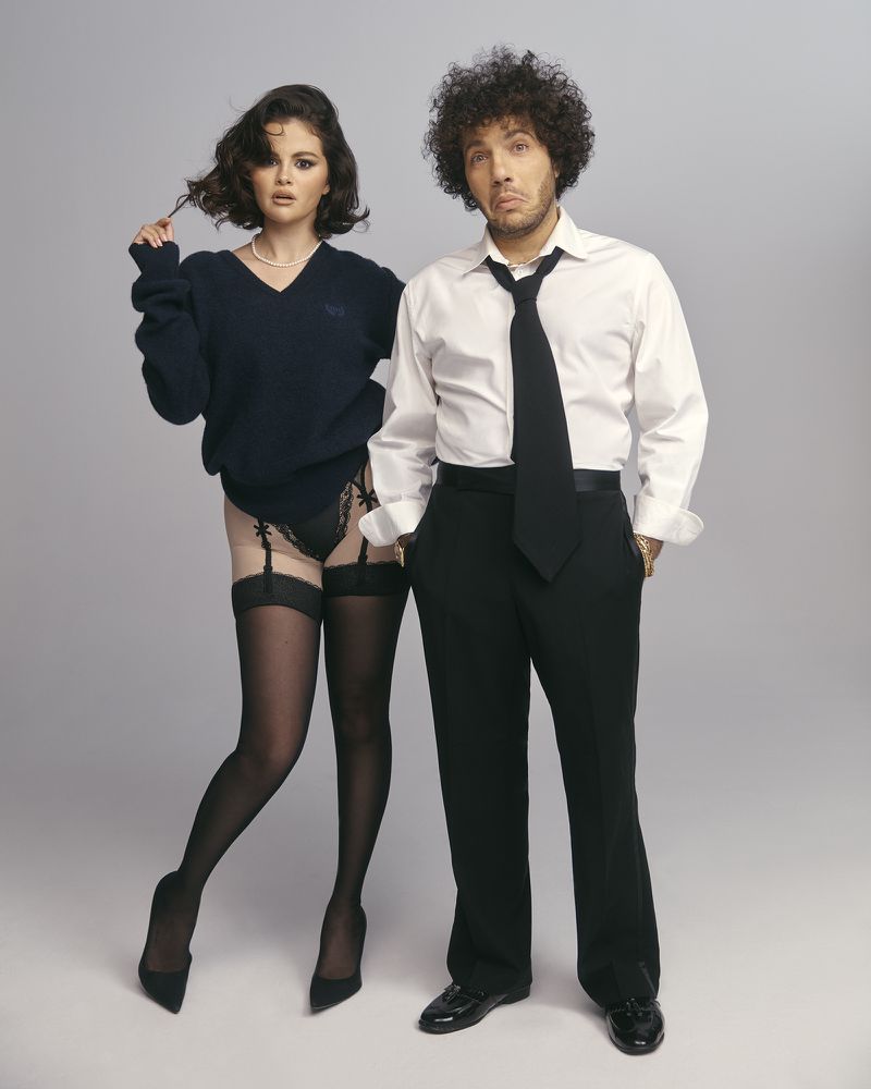 Selena Gomez and Benny Blanco pose together in a studio setting; she wears a sweater, black lingerie, and stockings, while he wears a suit.