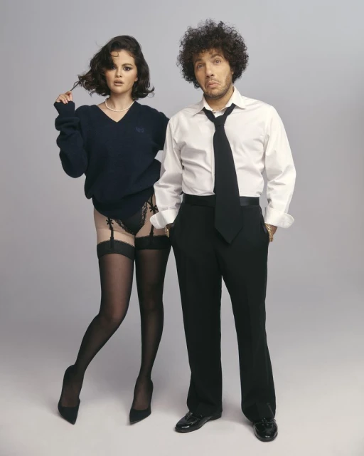 Selena Gomez and Benny Blanco pose together in a studio setting; she wears a sweater, black lingerie, and stockings, while he wears a suit.