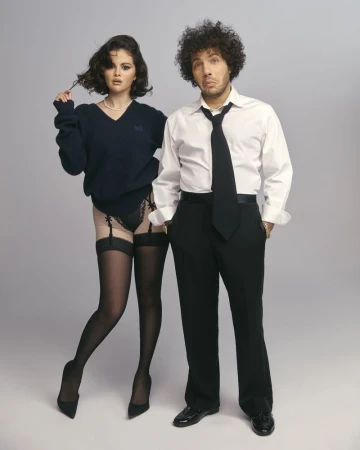 Selena Gomez and Benny Blanco pose together in a studio setting; she wears a sweater, black lingerie, and stockings, while he wears a suit.