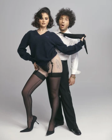 Selena Gomez and Benny Blanco pose in a studio setting. Gomez wears black sweater and stockings. Blanco wears shirt and trousers.