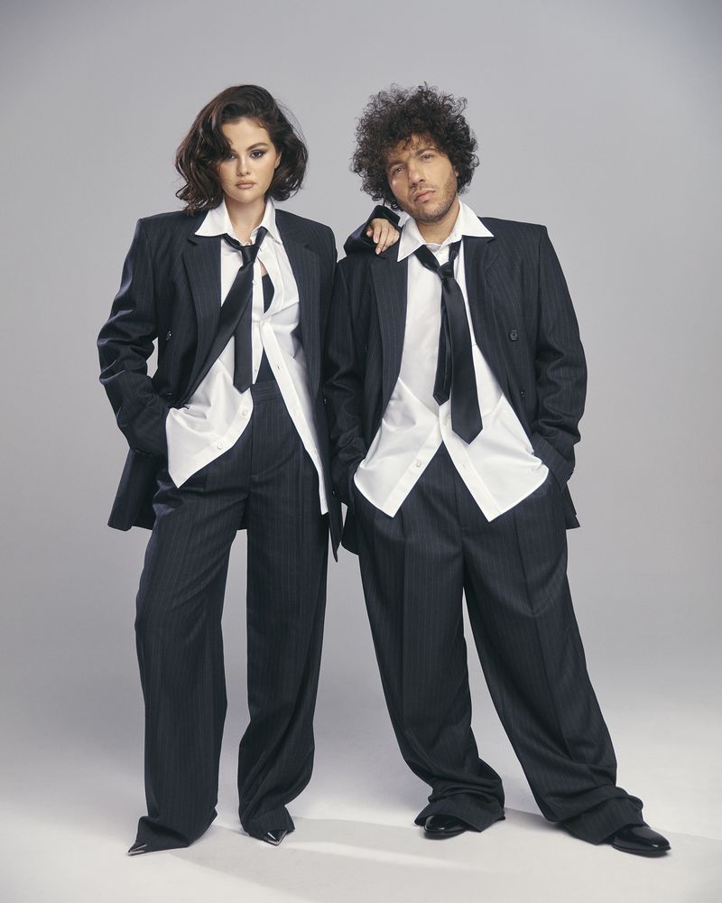 Two people wearing dark suits and light shirts stand in a photo studio, exuding a confident, stylish, and professional look.