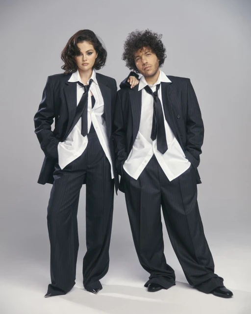 Two people wearing dark suits and light shirts stand in a photo studio, exuding a confident, stylish, and professional look.