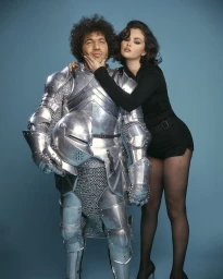 Benny Blanco in silver metal armor holding a helmet, with Selena Gomez standing beside him leaning on his shoulder in black attire.